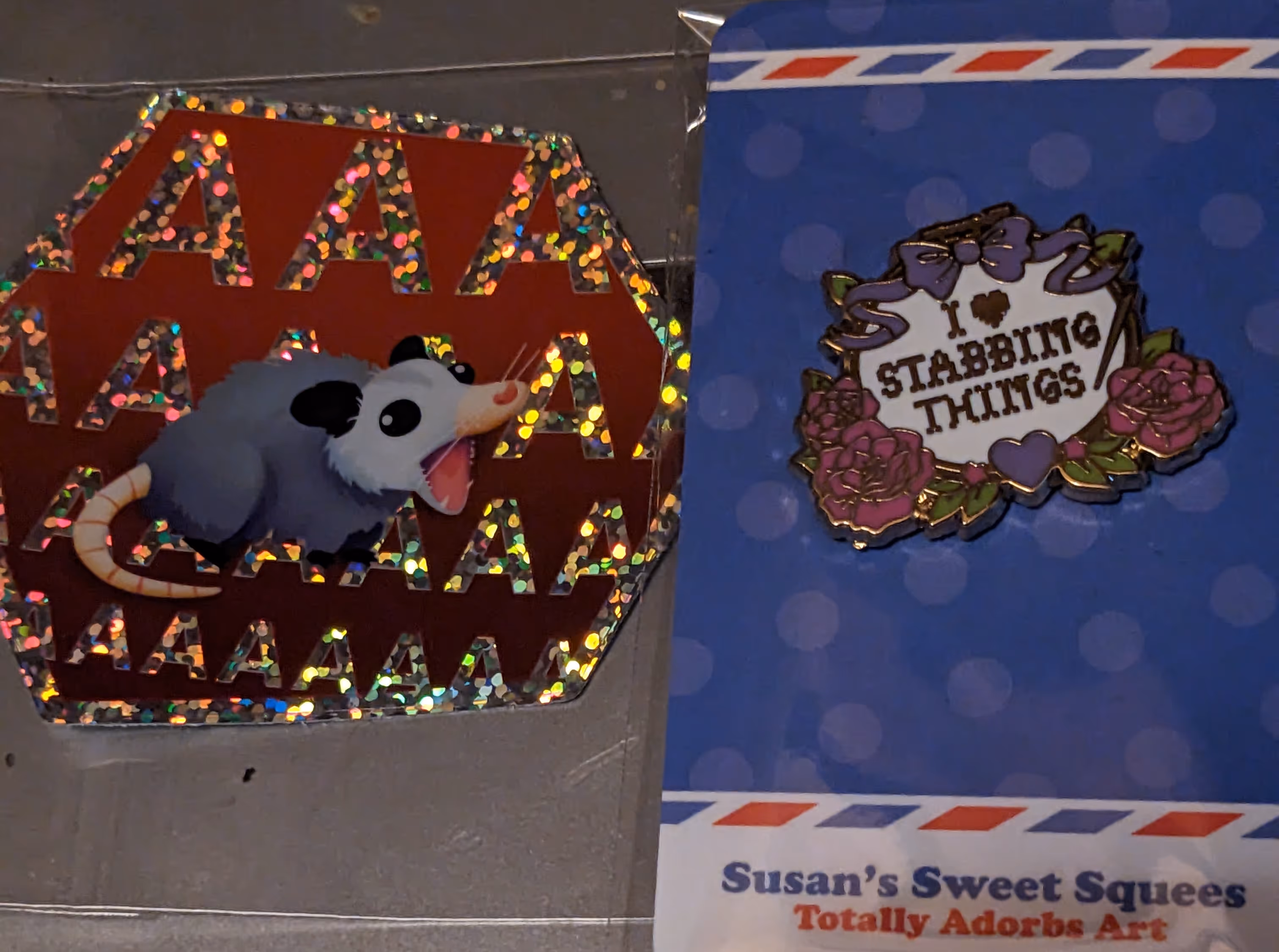 Image a sticker of a possum screaming and an enamel pin that says I heart stabbing things