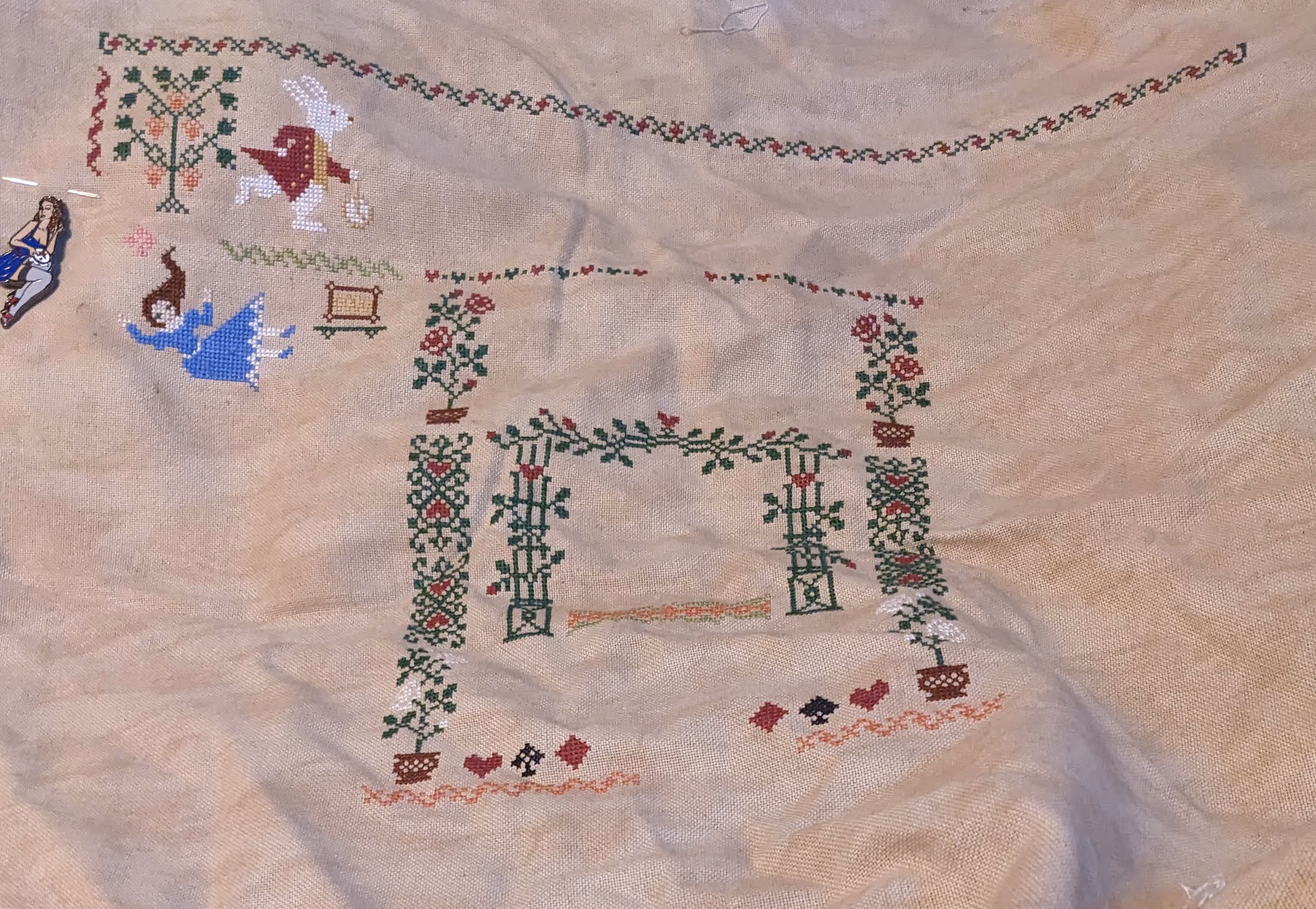 A large cross stitch featuring the white rabbit from Alice in Wonderland with several borders of roses and suits.