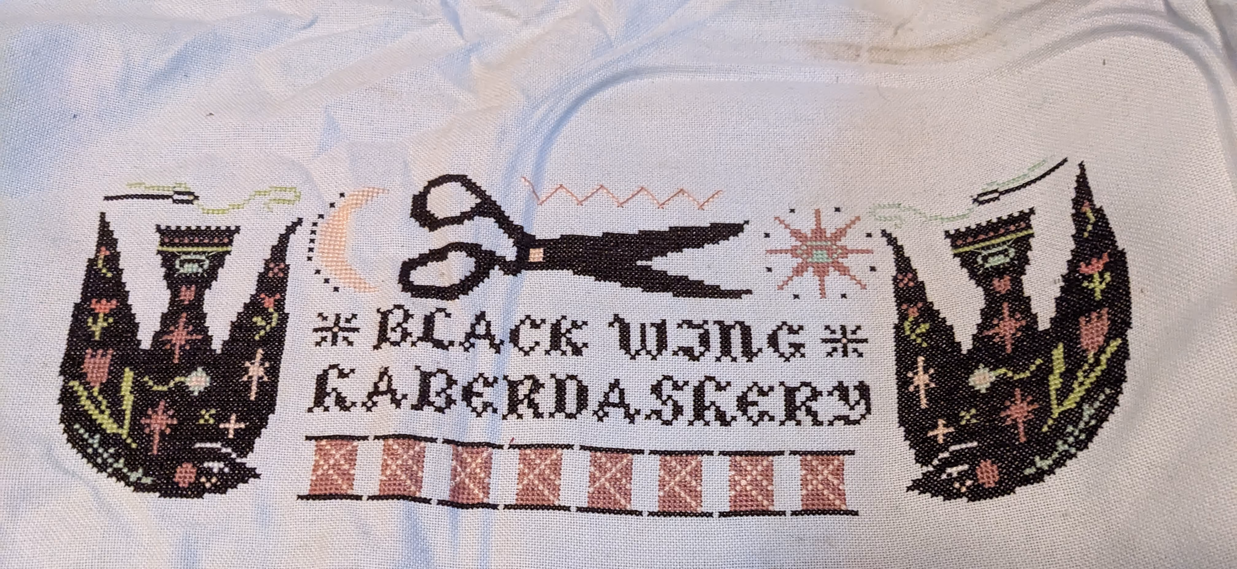 A cross stitch with the words black wing haberdashery and pink spools of thread along with a sun, moon, and threaded needles