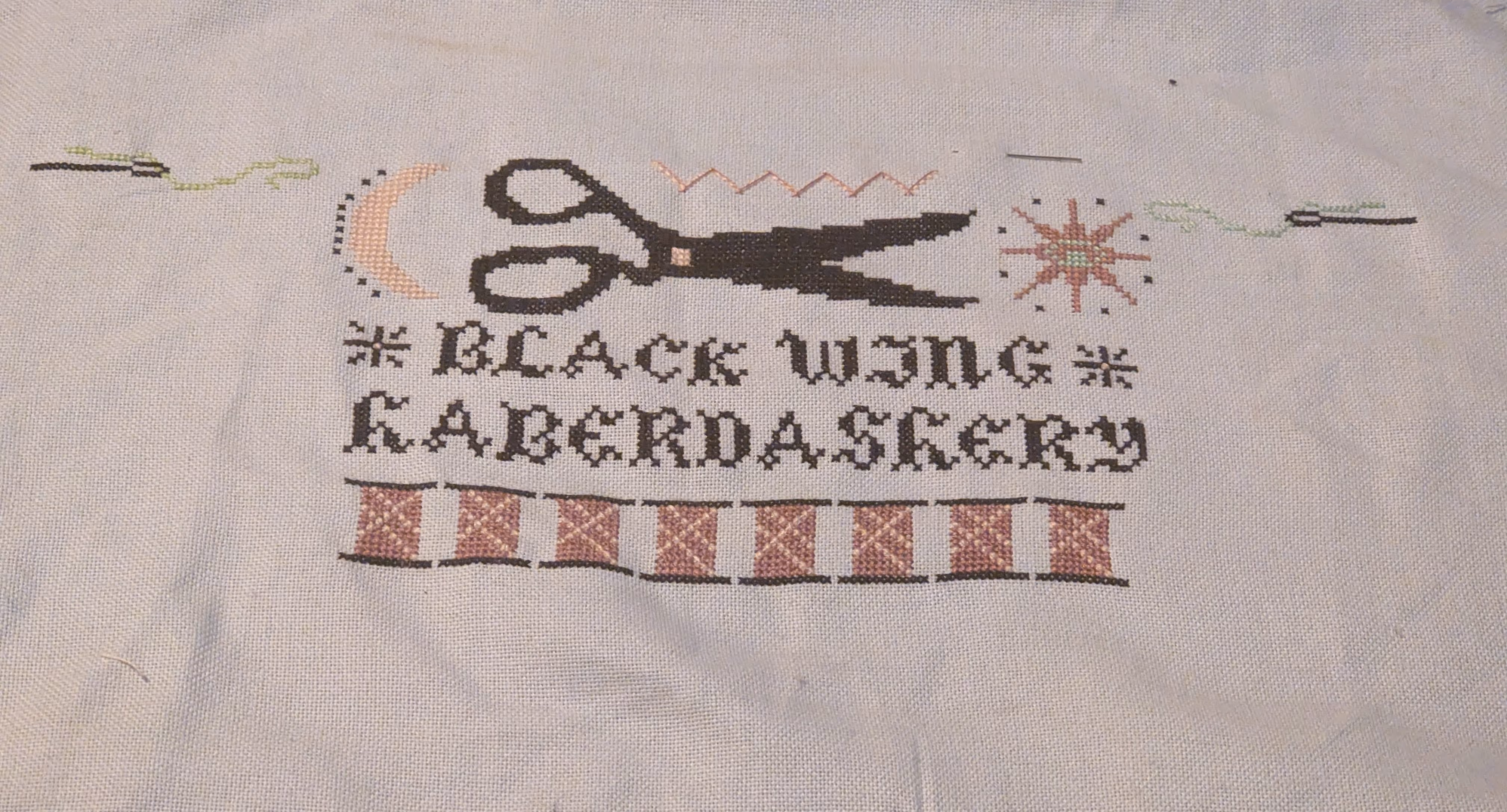 A partial cross stitch with the words black wing haberdashery and pink spools of thread along with a sun, moon, and threaded needles