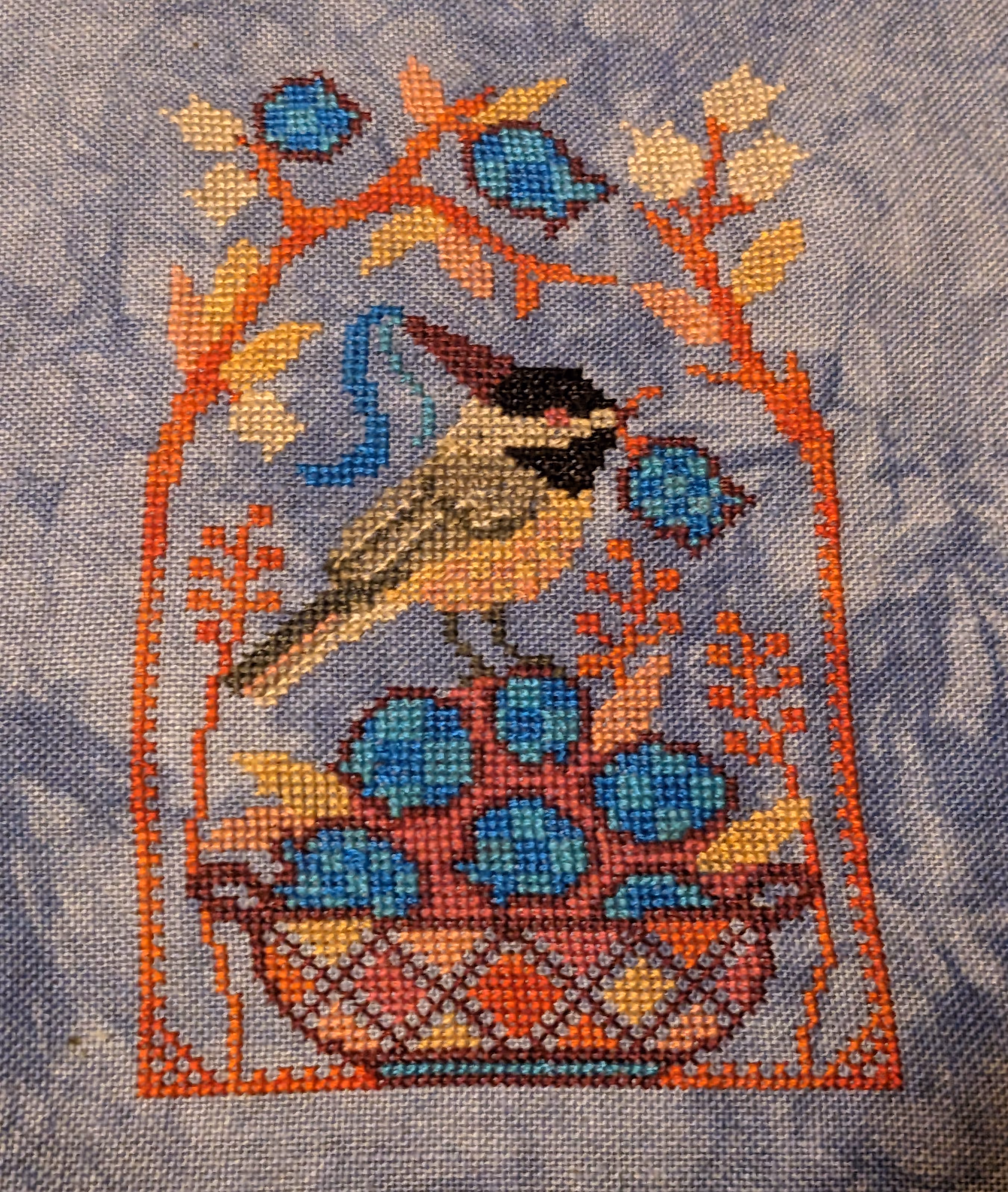 A chickadee in a princess hat has a blueberry in its mouth standing on a pile of blueberries surrounded by branches and flowers.