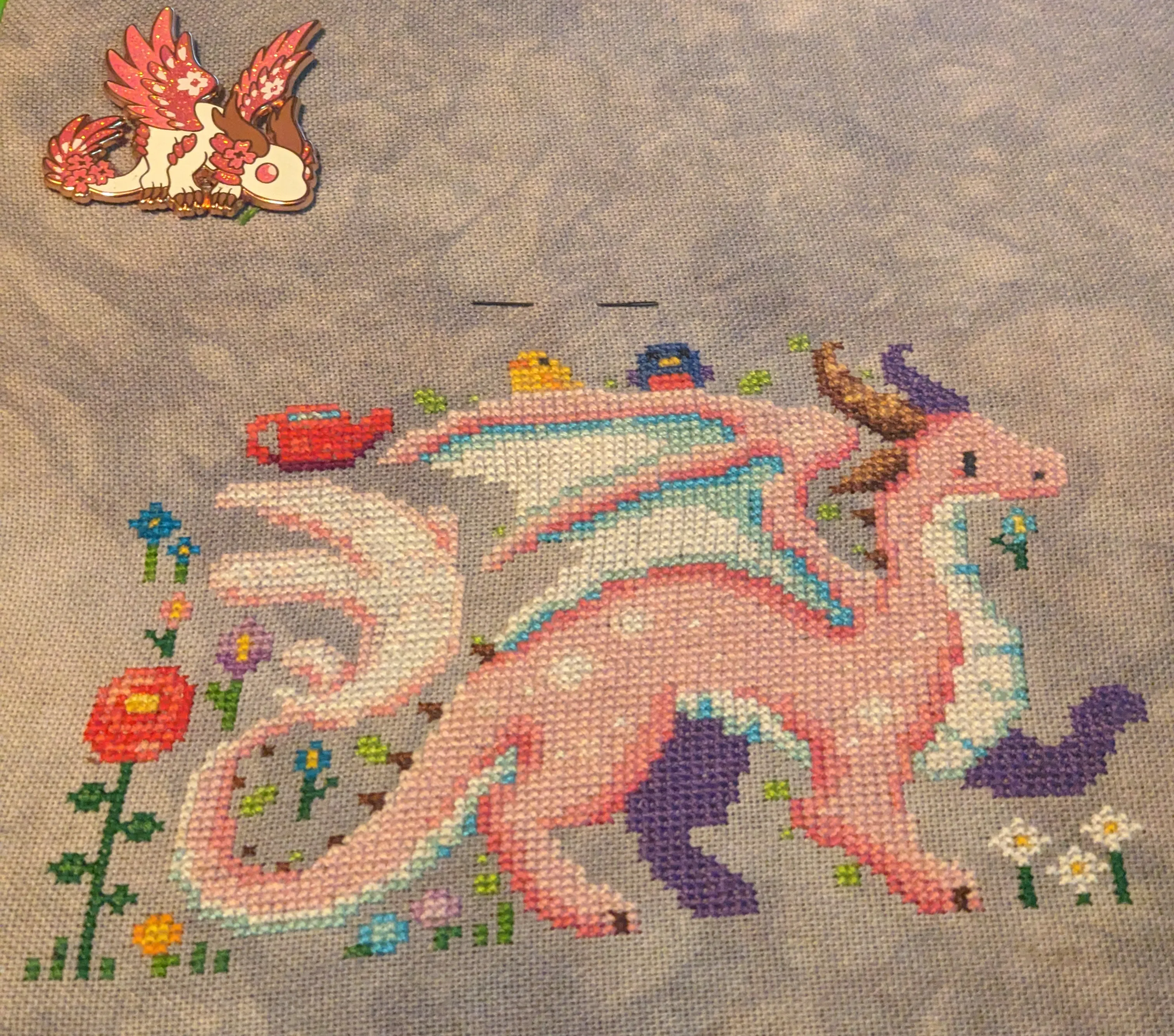 A cross stitch of a pink dragon with white spots in a meadow