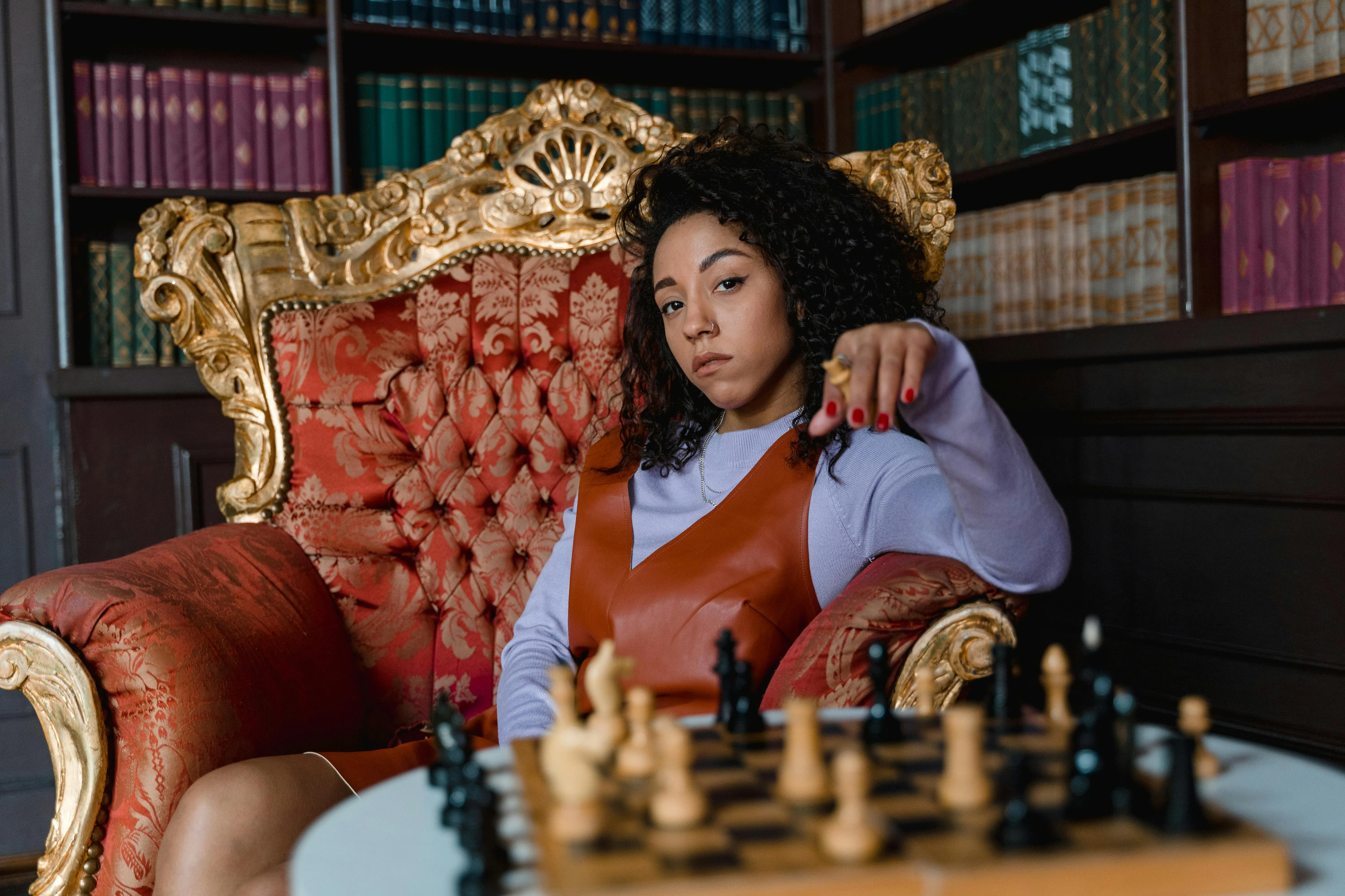 A woman lounges in a fancy chair holding a chess piece above a chess game on a table 