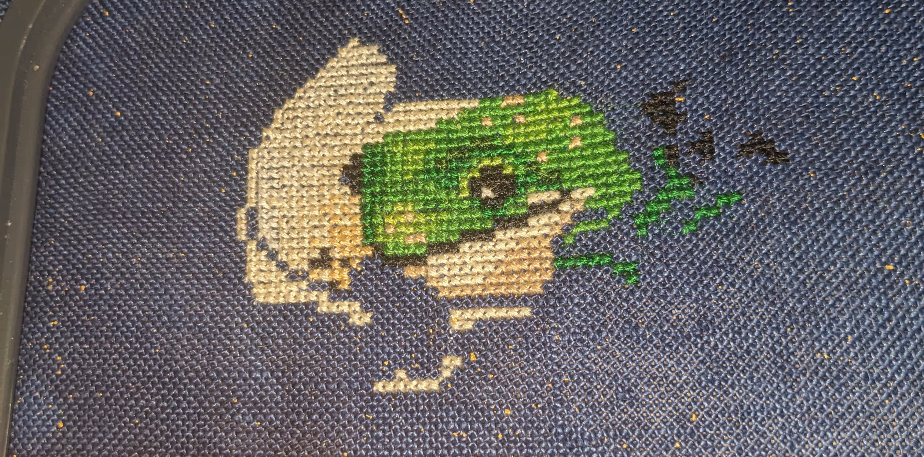 Image of a partial cross stitch showing a frog's face and the beginning of a tea cup.